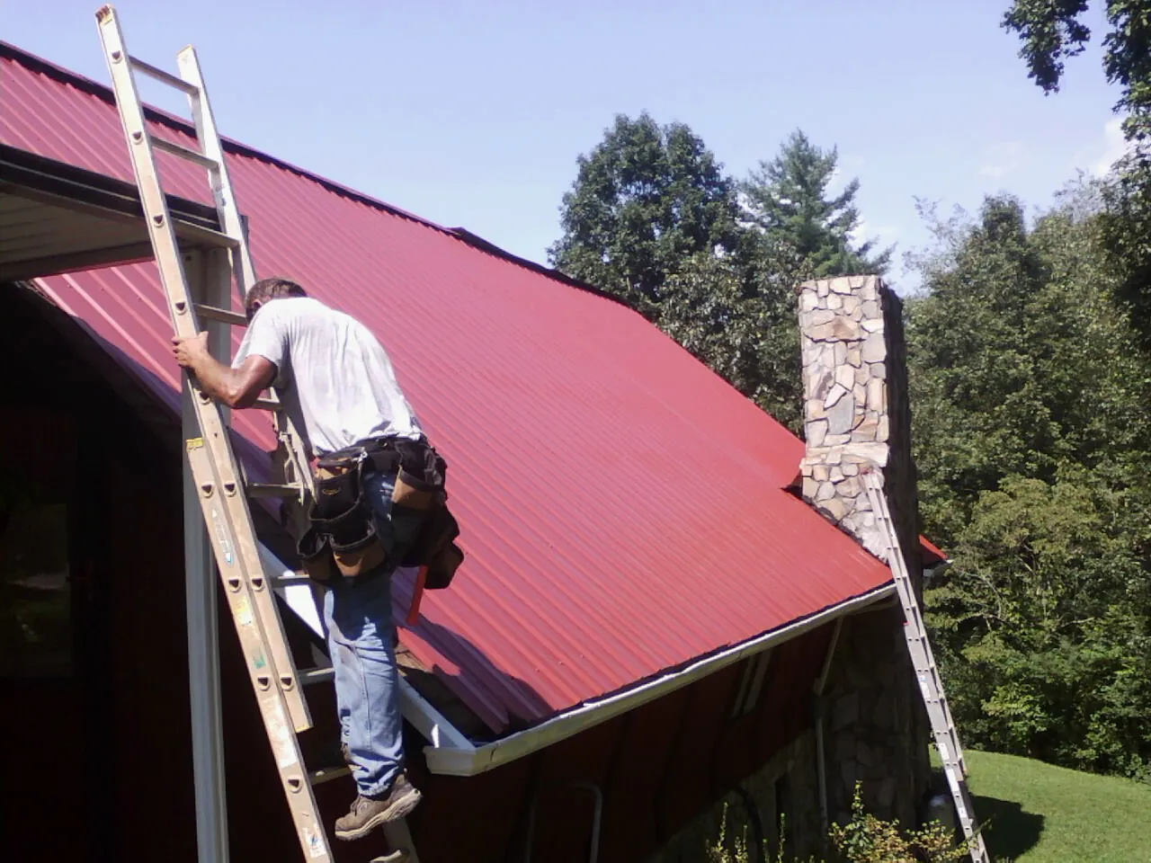 Roofer on a ladder beside a red metal roof and stone chimney — Soffit & Fascia Repair by Roofing Brothers in Desoto